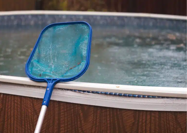Retino blu appoggiato al bordo di una piscina fuori terra con acqua sporca e detriti in superficie, utilizzato per la corretta pulizia della piscina